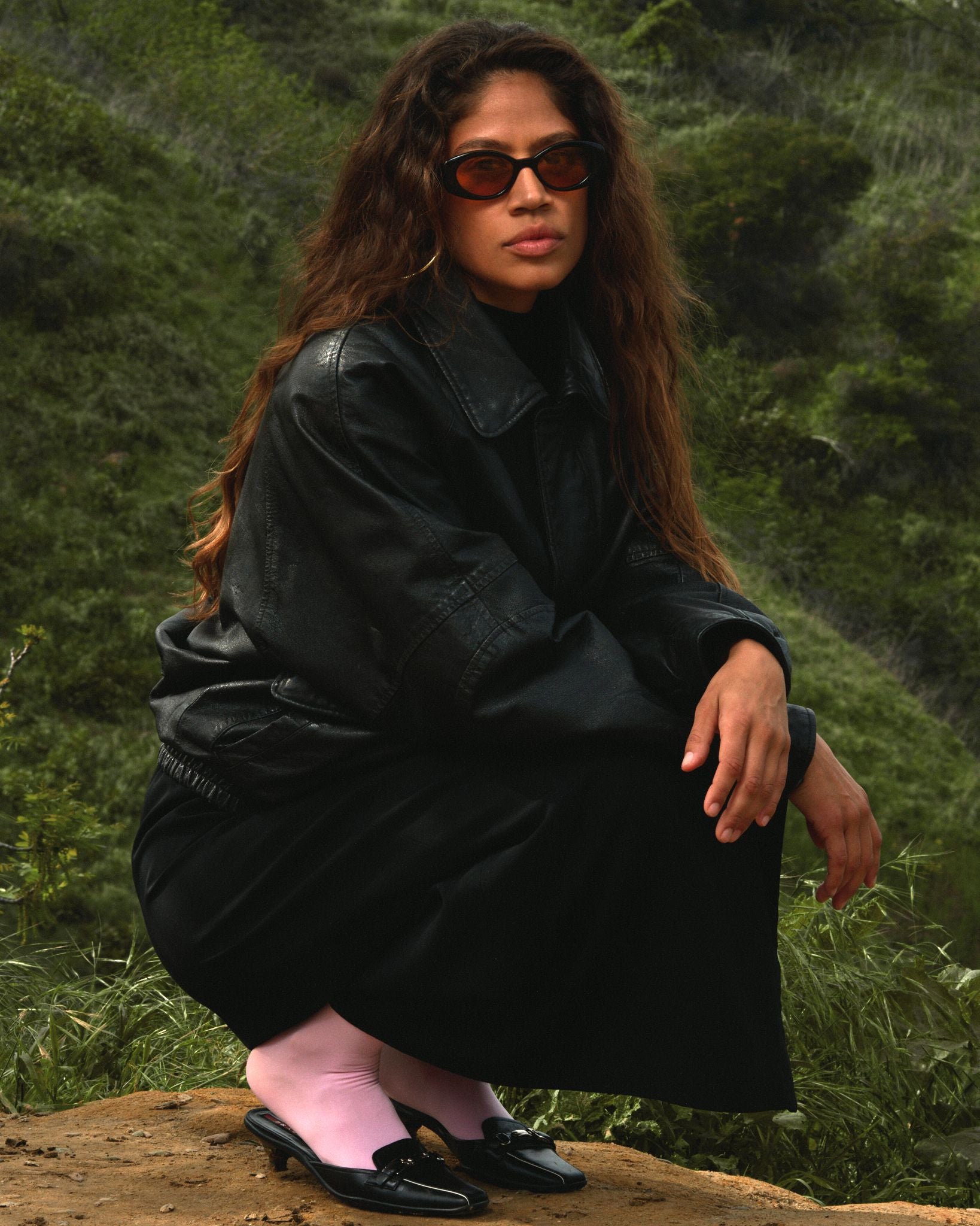 Beautiful ethnic girl wearing black framed sunglasses with an orange lens. Lush green scenery behind her.